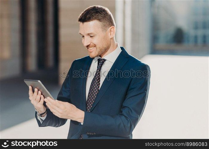 Cheerful male manager reads notification on digital tablet, wears formal black suit with tie, connected to wireless internet, watches video on web page. Business and modern technologies concept
