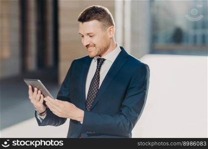 Cheerful male manager reads notification on digital tablet, wears formal black suit with tie, connected to wireless internet, watches video on web page. Business and modern technologies concept