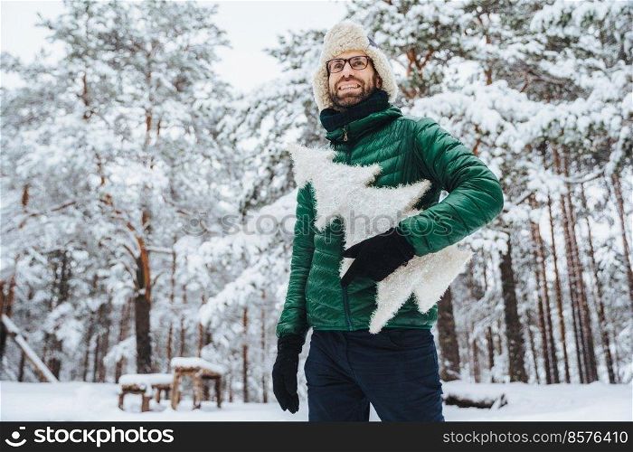 Cheerful male model dressed in winter clothes, holds white artificial fir tree, stands in winter forest, has pleasant smile, enjoys beautiful landscapes. People, holidays, recreation concept