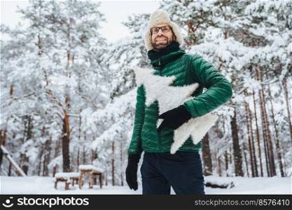 Cheerful male model dressed in winter clothes, holds white artificial fir tree, stands in winter forest, has pleasant smile, enjoys beautiful landscapes. People, holidays, recreation concept
