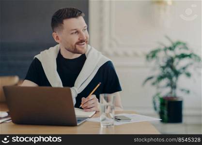 Cheerful male student searches information for homework via laptop writes article sits in coworking space alon looks away with happy smile on face watches training webinar creats personal blog