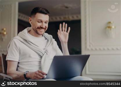 Cheerful man freelancer in wireless headphones wearing casual clothes waving to somebody and smiling while sitting in front of computer, having video call with colleagues at modern home office. Portrait of businessman or freelancer greeting somebody via laptop while sitting on chair at home