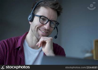 Cheerful man German speaking callcenter consultant in headset with mic works remotely from home, politely smiling while communicates online via laptop with customers, helps in solving current problems. Cheerful man German speaking call center consultant in headset with mic works remotely from home