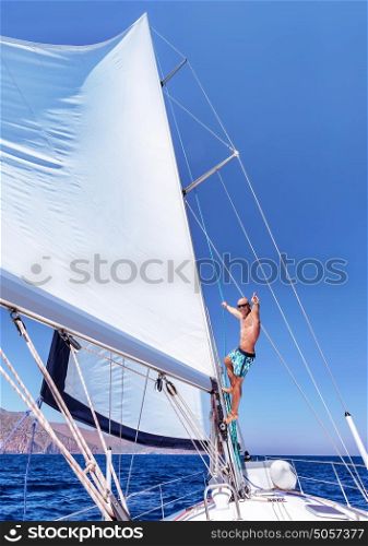 Cheerful man having fun on sailboat, sitting up on the mast of water transport, enjoying active summer vacation, enjoyment and happiness concept
