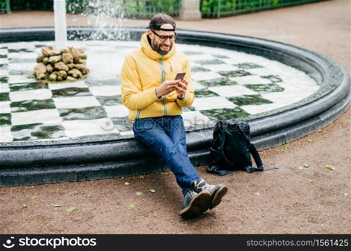 Cheerful man in cap, eyeglasses and yellow anorak sitting near fountain chatting with friends using mobile phone. Tired man after walk in big city having rest near fountain watching video online