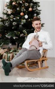 cheerful man sitting on sleigh at home near Christmas tree. man on a sleigh. Christmas good mood. Family and holiday concept. cheerful man sitting on sleigh at home near Christmas tree. man on a sleigh. Christmas good mood. Family and holiday concept.