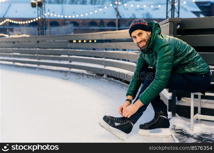 Cheerful man with appealing appearance laces up skates, sitts on ice arena, wants to go skating with friends, poses at camera. Happy bearded man enjoys having active lifestyle. Winter and season