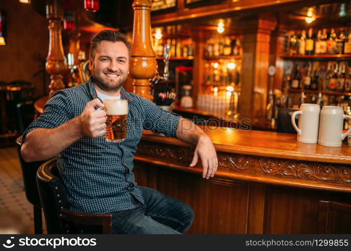 Cheerful man with beer mug at the counter in pub. Bearded male person with glass of alcohol having fun in bar