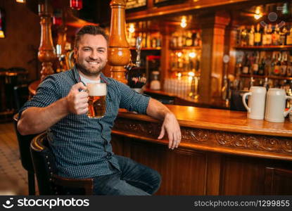 Cheerful man with beer mug at the counter in pub. Bearded male person with glass of alcohol having fun in bar