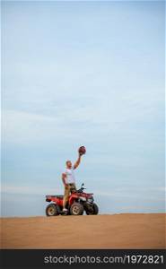 Cheerful man with helmet in hand poses on atv, downhill riding in desert sands. Male person on quad bike, sandy race, dune safari in hot sunny day, 4x4 extreme adventure, quad-biking. Man with helmet in hand poses on atv in desert
