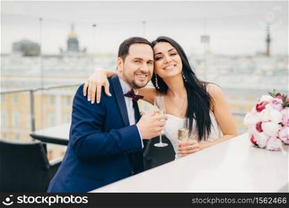Cheerful married couple embrace each other, clink glasses with champagne, have good mood after registring their marriage, celebrate alone, wait for guests, pose at camera with pleased smiles