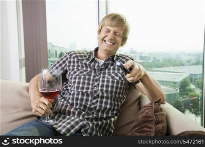 Cheerful mid-adult man with wine glass watching television on sofa at home