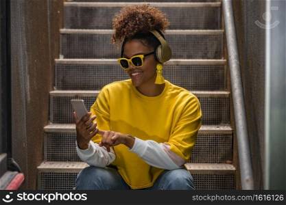 Cheerful millennial African American female in trendy yellow sunglasses and wireless headphones choosing playlist on mobile phone while sitting on staircase. Trendy black hipster woman using smartphone
