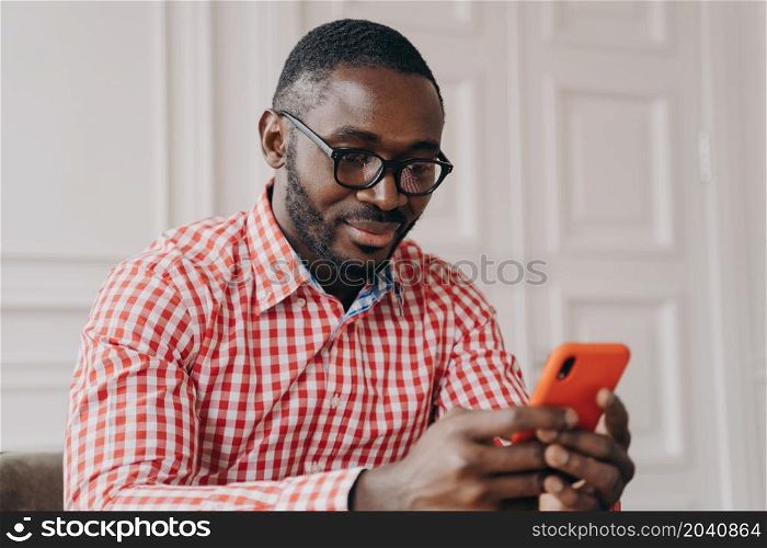 Cheerful millennial biracial man using smartphone, sitting at desk , young Afro american guy carefully looking on phone screen while typing urgent message, chatting online or searching in internet. Cheerful millennial biracial man using smartphone, sitting at desk