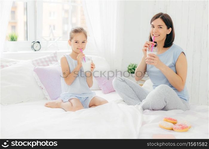 Cheerful mother and daughter dressed in pyjamas, have breakfast in morning, drink milk shake with doughnuts, sit crossed legs on comfortable bed in white bedroom. People and lifestyle concept