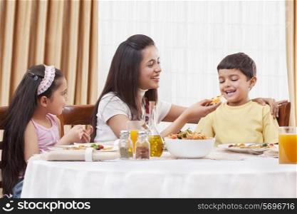 Cheerful mother feeding her son pizza with daughter sitting besides