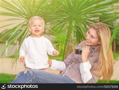 Cheerful mother taking picture of her cute little daughter in the park, happy young family having fun outdoors, parental love and enjoyment