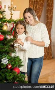 Cheerful mother with daughter decorating Christmas tree with colorful baubles