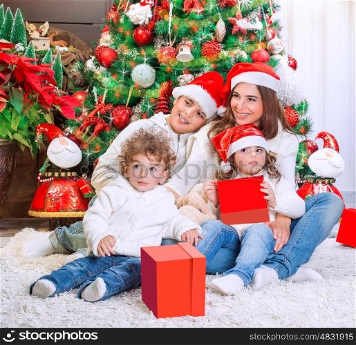 Cheerful mother with her three precious kids wearing red Santa hats sitting near beautiful decorated Christmas tree, happy family holiday