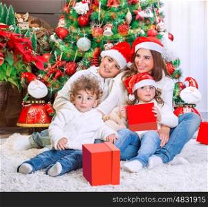Cheerful mother with her three precious kids wearing red Santa hats sitting near beautiful decorated Christmas tree, happy family holiday