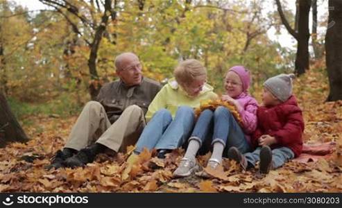 Cheerful multi-generation family spending freetime in public park on fall day. Portrait of positive grandparents and lovely grandchildren sitting on fallen leaves and relaxing outdoors in autumn during indian summer. Slow motion.