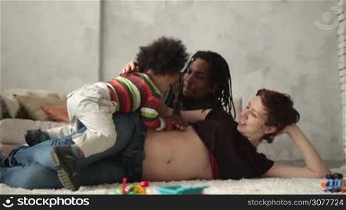 Cheerful multiethnic family spending leisure together at home. Caucasian pregnant mother lying on carpet, african american father with dreadlocks touching belly and kissing wife&acute;s shoulder while curly mixed race toddler son playing. Slow motion.