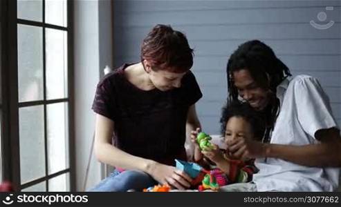 Cheerful multiethnic parents and curly little mixed race toddler son playing with colorful toys at home. Positive african american father, joyful caucasian mother and their cute boy having fun in the bedroom while spending good time together.