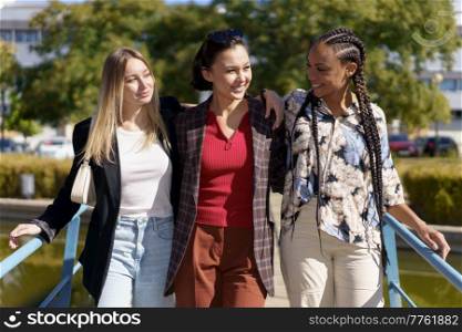 Cheerful multiracial female friends looking at each other while standing together on bridge over river in city on summer day. Positive diverse women on footbridge in city