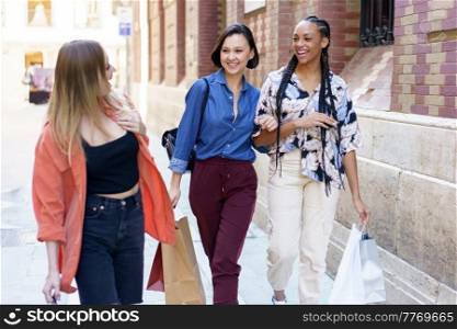 Cheerful multiracial female friends with shopping bags looking at each other while strolling together on walkway near building in city. Smiling diverse women with shopping bags walking on street