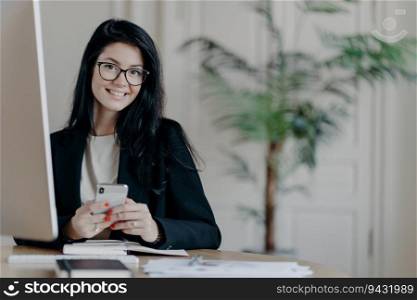 Cheerful office worker, stylish in black jacket, uses smartphone happily while working on an important project in a modern coworking space. Modern technologies.