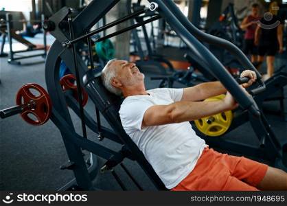 Cheerful old man on exercise machine, gym interior on background. Sportive grandpa on fitness training in sport center. Healty lifestyle, health care. Cheerful old man on exercise machine in gym