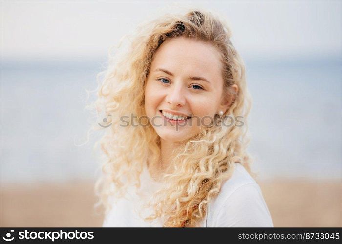 Cheerful optimistic young female has shining smile, curly hair, breathes fresh air at seaside, stands alone, has fresh skin. Attractive woman enjoys leisure time, expresses happiness. Emotions concept