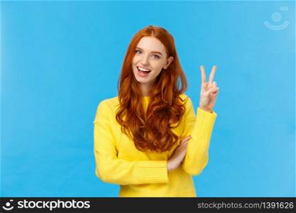 Cheerful outgoing and happy charismatic redhead female in yellow sweater, showing peace sign, number two or twice and smiling, making purchase, order double cheese, blue background.. Cheerful outgoing and happy charismatic redhead female in yellow sweater, showing peace sign, number two or twice and smiling, making purchase, order double cheese, blue background