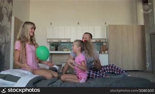 Cheerful parents and adorable blonde daughter playing together with balloon on bed in the morning. Carefree family spending free time with their little girl and having fun while relaxing at home over modern room background. Slow motion. Dolly shot.