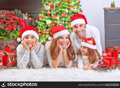 Cheerful parents with two cute smiling kids lying down near Xmas tree at home, celebrating Christmas, festive greeting card, happy family holiday concept