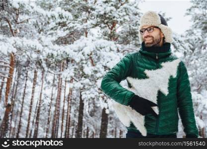 Cheerful pleasant looking man with beard looks thoughtfully aside, holds fir tree, wears spectacles, hat and jacket, poses against winter trees covered with snow. People and lifestyle concept