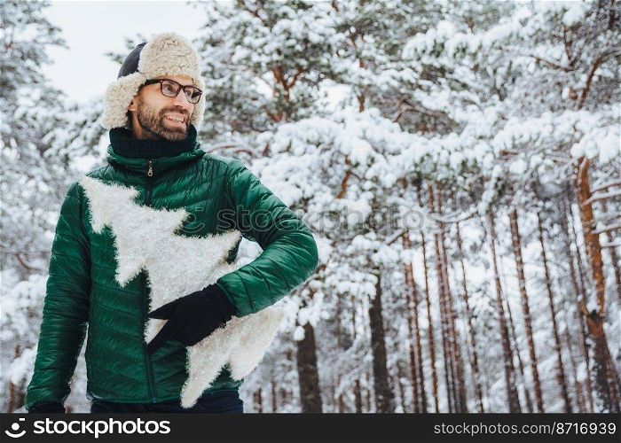 Cheerful pleasant looking man with beard looks thoughtfully aside, holds fir tree, wears spectacles, hat and jacket, poses against winter trees covered with snow. People and lifestyle concept