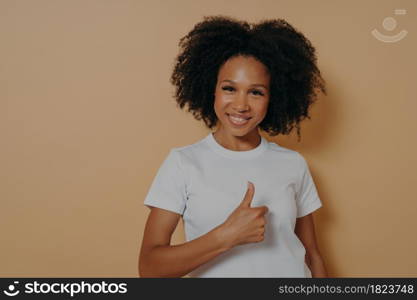 Cheerful positive young african woman showing success gesture, thumb up with hand and smiling at camera with positive face expression, saying well done, isolated on beige studio background. Cheerful positive young african woman saying well done, showing thumb up while standing in studio
