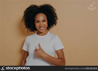 Cheerful positive young african woman showing success gesture, thumb up with hand and smiling at camera with positive face expression, saying well done, isolated on beige studio background. Cheerful positive young african woman saying well done, showing thumb up while standing in studio