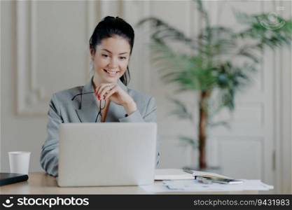 Cheerful professional woman searches laptop update, removes glasses in formal office attire, working remotely on blog ideas.