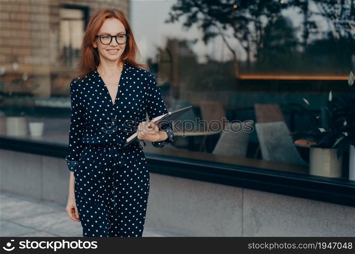 Cheerful prosperous businesswoman in elegant outfit holds tablet and diary looks happily into distance waits for partner of business meeting stands outdoor near restaurant strolls in urban place. Cheerful prosperous businesswoman in elegant outfit holds tablet and diar
