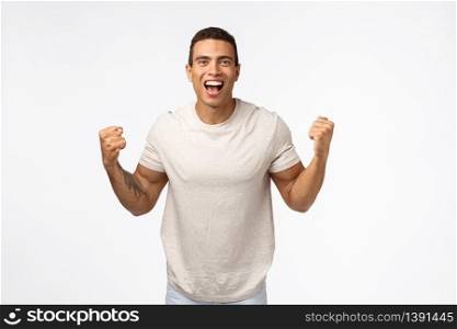 Cheerful, proud attractive young hispanic man with tattoo arm, clench fists and yelling upbeat, achieve goal, winning lottery or competition, become champion, say yes or hooray white background.. Cheerful, proud attractive young hispanic man with tattoo arm, clench fists and yelling upbeat, achieve goal, winning lottery or competition, become champion, say yes or hooray white background