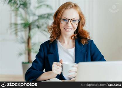 Cheerful redhead woman prepares application letter, sits in front of opened laptop computer, browses internet and seeks for job vacancies, holds mug with hot drink, wears spectacles and formal outfit