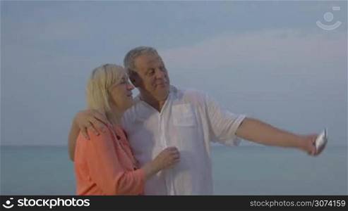 Cheerful senior couple on vacation taking selfie with smartphone. Picture in hugs of each other on sea background