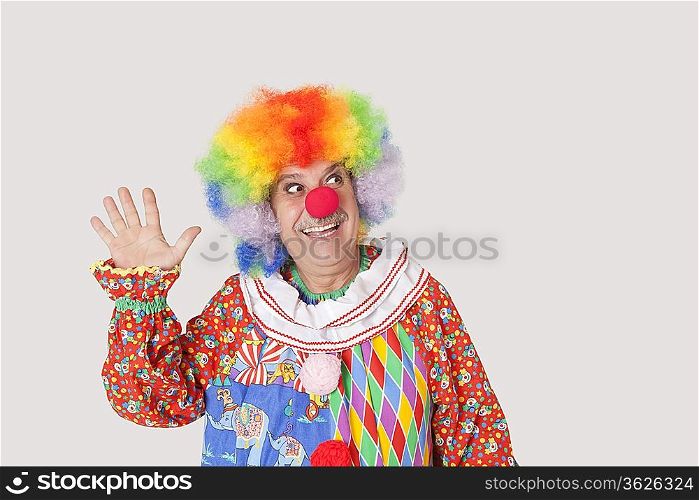 Cheerful senior male clown waving hand while looking away over gray background