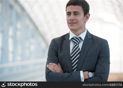 Cheerful serious young businessman standing in urban surrounding with arms crossed