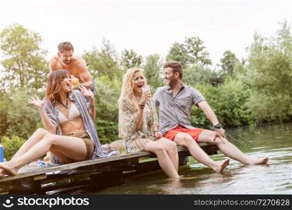 Cheerful shirtless man spraying water with squirt gun on friends sitting at pier over lake