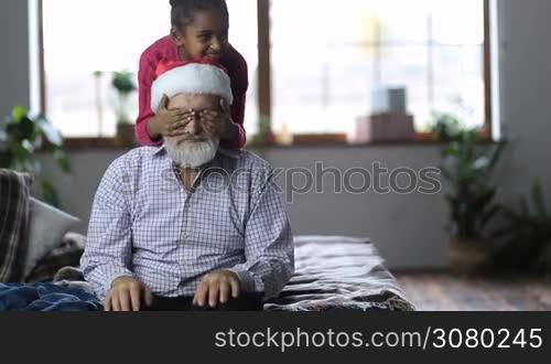 Cheerful small mixed race girl holding eyes of her grandfather closed as her younger sister giving christmas gift box to grandfather. Joyful grandpa in sant hat with white beard receiving xmas present from adorable granddaughters on winter holidays.