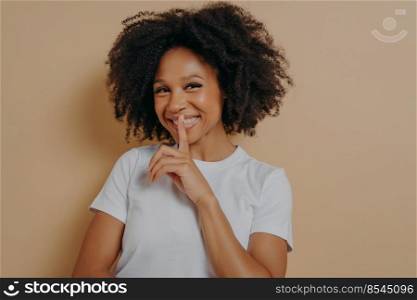 Cheerful smiling afro girl showing shhh sign, keep silence gesture, with index finger near lips, standing over pastel beige background and looking at camera. Positive emotions and body language. Cheerful smiling afro girl showing shhh sign, keep silence gesture, with index finger near lips