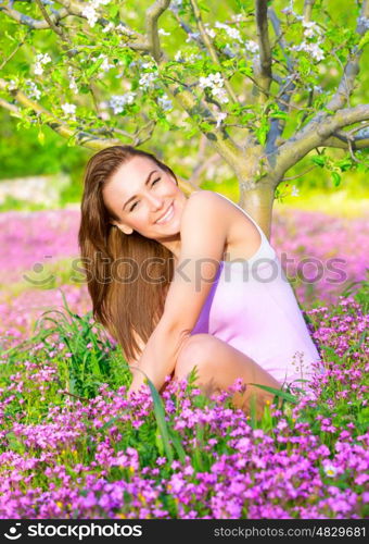 Cheerful smiling female spending time on backyard, enjoying apple tree blossom, sitting on pink floral glade, spring season concept